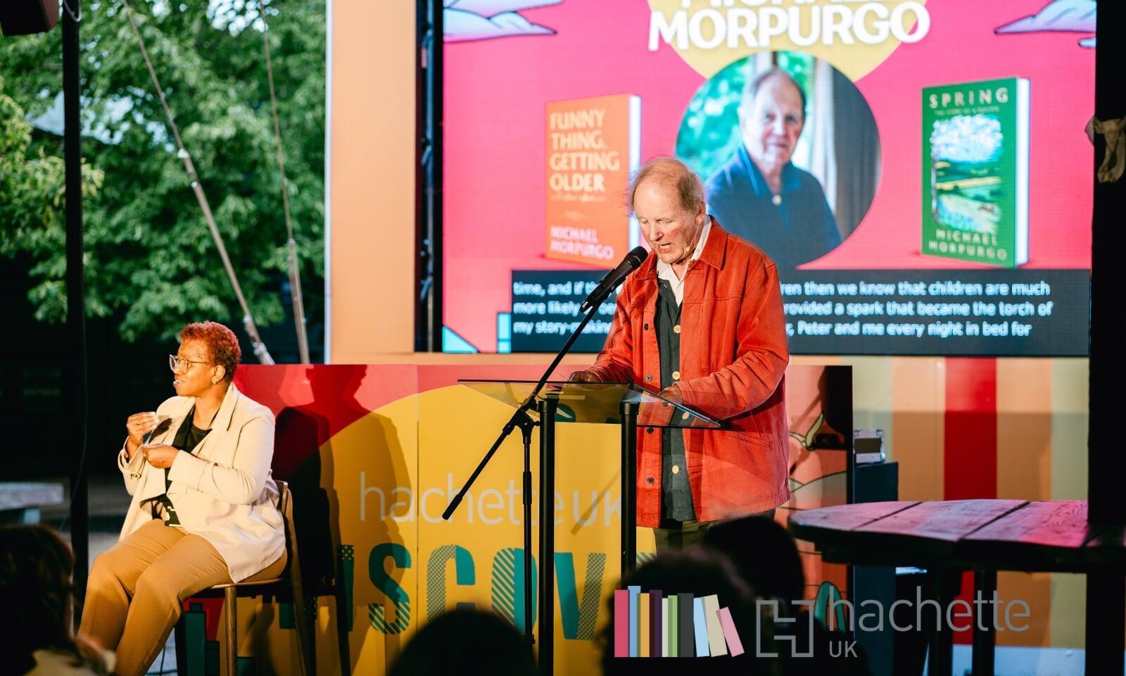 An image of a brightly coloured stage with an author reading from his book and a BSL interpreter sitting on a chair to his right